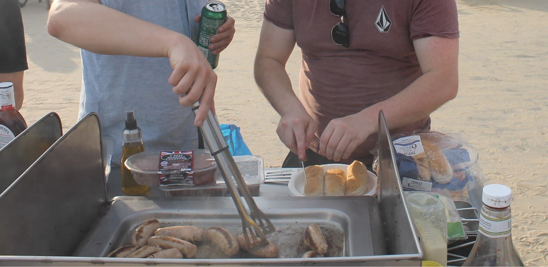 Two people cooking sausages on a stainless-steel electric grill set on sand, with tongs, bread rolls and a drinks can nearby