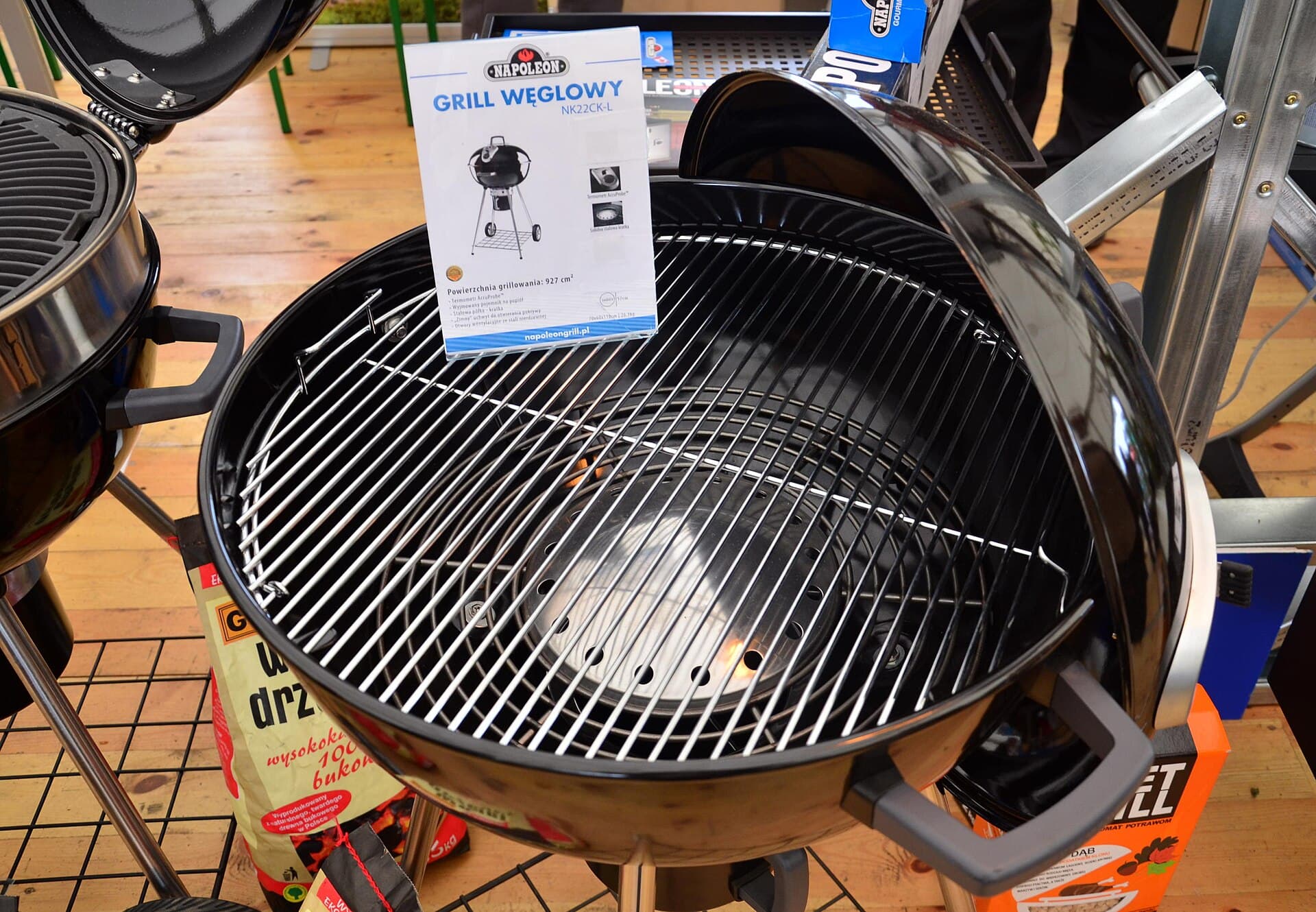 A black kettle charcoal barbecue with its lid open and a chrome grate, displayed on a timber floor beside a product information sheet