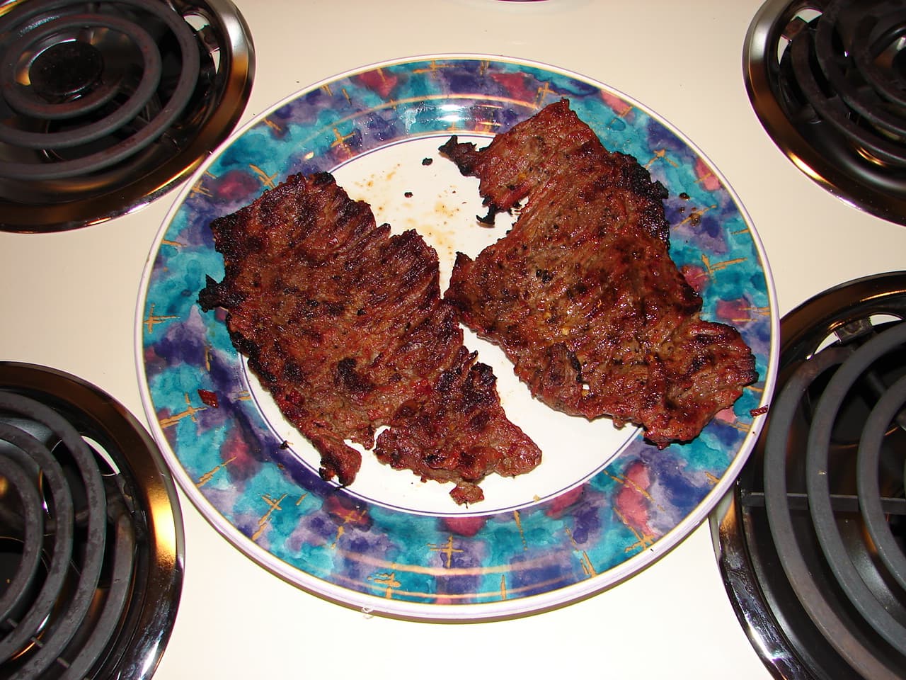 Two pieces of grilled skirt steak on a patterned plate sitting on a white electric cooktop