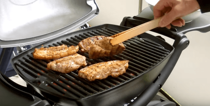 A compact black portable barbecue cooking several pieces of meat on a ridged grate, with wooden tongs lifting one piece